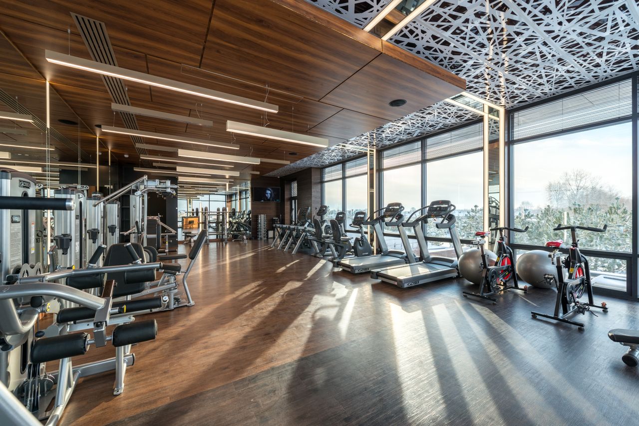 Modern exercise bikes and treadmills and various fitness equipment in spacious gym with wooden floor and panoramic windows in sunlight
